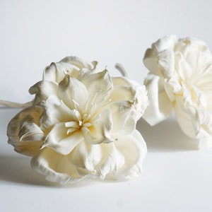 Op de afbeelding: Twee ivoorkleurige kunstbloemen met gedetailleerde bloemblaadjes. Eén bloem is volledig open, met een gele kern, terwijl de andere gedeeltelijk open is. De bloemen staan tegen een witte achtergrond, wat wijst op een bloemen- of decoratiethema.