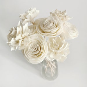 Op de afbeelding: Een boeket witte sola-houten bloemen in een heldere glazen vaas. De bloemen zijn in een cirkelvormige opstelling geplaatst en hebben een delicate, natuurlijke uitstraling.