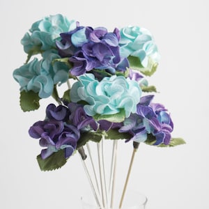 May include: A bouquet of artificial hydrangeas in shades of blue and purple. The flowers have layered petals and green leaves. The stems are thin and white, and the arrangement is in a clear glass vase.
