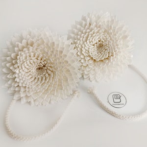 Op de afbeelding: Twee grote witte papieren bloemen met een touwlus aan elke. De bloemen zijn gemaakt van vele lagen papier en hebben een delicate, gelaagde uitstraling.