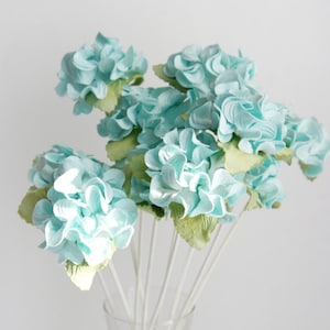 Peut inclure: Un bouquet d'hydrangeas artificiels bleu clair avec des feuilles vertes. Les fleurs sont regroupées sur des tiges blanches, créant un aspect plein et texturé. L'arrangement est posé sur un fond blanc uni.