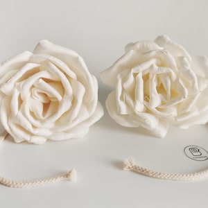 Op de afbeelding: Twee witte sola-houten rozen met een wit katoenen koord aan elke roos bevestigd. De rozen zijn symmetrisch gerangschikt.