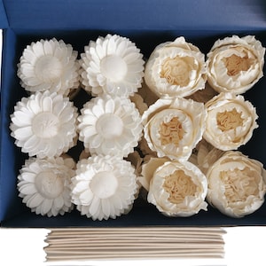 May include: A blue box filled with white sola wood flowers. The flowers are arranged in two rows, with the top row containing a mix of sunflower and peony shapes. The bottom row contains only sunflower shapes. The box is open and the flowers are visible. There are also several wooden slats stacked on top of the box.