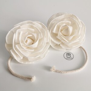 May include: Two white sola wood flower rosettes with a white cotton cord. The flowers are arranged in a spiral pattern and have a delicate, ruffled texture. The cord is tied around the flowers and hangs down.