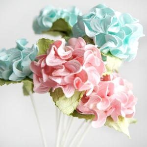 May include: A collection of handmade paper flowers in pink and teal hues. The flowers have a layered, textured appearance, with green paper leaves. The stems are white. The flowers are arranged against a white backdrop.