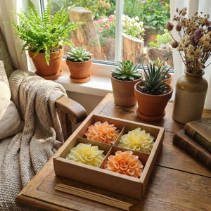May include: A square cardboard box containing four flower-shaped wax candles in shades of peach and pale yellow, arranged in a grid. The candles feature intricate, layered petal designs. A bundle of thin wooden sticks lies on the table in front of the box.