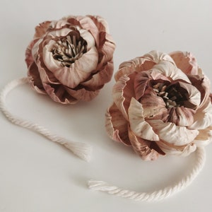 Op de afbeelding: Twee beige en bruine sola-houten bloemstukken met een witte touwtje eraan bevestigd. De bloemen zijn in een pioenroos-stijl.