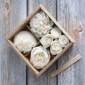 May include: A collection of white, decorative flowers in a divided cardboard box. The assortment includes large, layered blooms and smaller rose-like buds. Several wooden diffuser sticks are placed beside the box, suggesting a fragrance product.