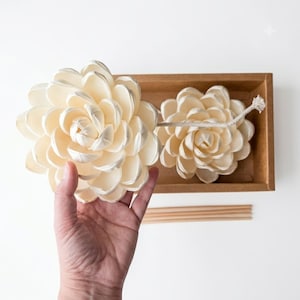 Puede incluir: Dos difusores en forma de flor de color crema, con múltiples capas de material fino y flexible. Una flor está unida a un cordón de algodón largo y fino, mientras que la otra descansa dentro de una caja de cartón marrón con palitos de madera.