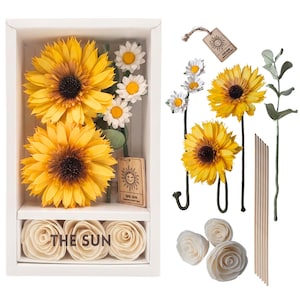 May include: A white rectangular box containing two large yellow sunflowers, three small white daisies, and three cream-colored roses with the text THE SUN printed on the front. Included are loose flower stems, wooden sticks, and a sun-themed tag.