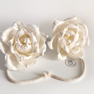 May include: Two white sola wood flower heads with a white cotton cord. The flowers are tied together with the cord. The flowers are in the shape of roses.