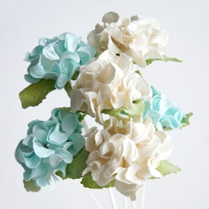 May include: A bouquet of artificial flowers featuring light blue and cream-colored blossoms with green leaves. The flowers have a layered petal design and are arranged on thin, white stems, creating a delicate and elegant floral arrangement.