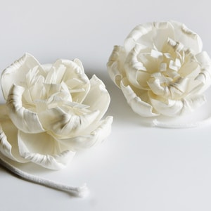 Op de afbeelding: Twee witte sola-houten bloemstukken met een touwtje aan elk. De bloemen zijn gemaakt van sola-hout en hebben de vorm van rozen.