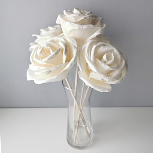 May include: A bouquet of four white paper roses in a clear glass vase with wooden stems.