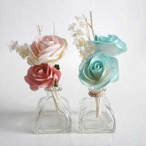 Op de afbeelding: Twee glazen flessen met vierkante bodems en heldere glazen deksels. Elke fles bevat een boeket kunstbloemen. Een boeket is roze en wit, de andere is blauw en wit. Beide boeketten hebben houten stokjes die zich uitstrekken van de bloemen in de flessen.