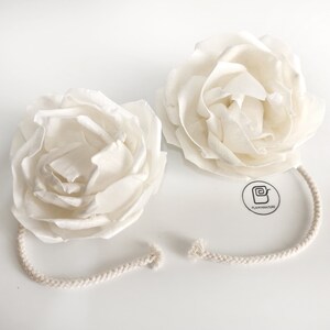 May include: Two white paper flower decorations with a white cotton rope attached to each. The flowers are made of paper and have a delicate, layered design. The rope is tied together in a knot.