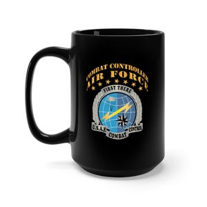 Może przedstawiać: Czarny ceramiczny kubek z dużym uchwytem. Kubek zawiera napis "COMBAT CONTROLLER AIR FORCE" złotymi literami, a także grafikę przedstawiającą globus, spadochron i gwiazdę. Wyświetlane są również napisy "FIRST THERE" i "U.S.A.F. COMBAT CONTROL".