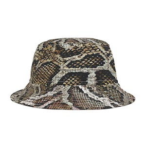 Puede incluir: Un sombrero de pescador con un estampado de piel de serpiente en tonos marrón, negro y blanco. El sombrero tiene una corona redondeada y un ala inclinada hacia abajo. La tela parece ser un textil estampado.