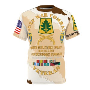 May include: A beige and white t-shirt featuring a military design. The shirt includes the text "GULF WAR COMBAT VETERAN" and "800th Military Police Brigade". The design incorporates an American flag, crossed pistols, and military insignia.