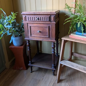 May include: A dark brown wooden side table featuring a rectangular top, one small upper drawer with vertical slats, and one larger lower drawer. The table stands on four turned legs with a lower shelf and small castors at the base of each leg.