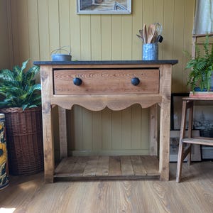 May include: A wooden console table featuring a dark grey stone top, a single wide drawer with two black round knobs, and a scalloped apron detail. The table includes a lower slatted shelf and is constructed from light brown natural wood.