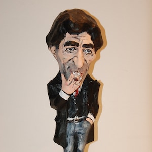 Puede incluir: Una figurita de papel maché hecha a mano de una persona que lleva un traje negro, una camisa blanca y una corbata roja. La figurita tiene una cara realista con bigote y sostiene un cigarrillo en la mano.