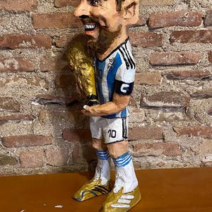 May include: A paper mache figurine of a soccer player holding a gold trophy. The player is wearing a blue and white jersey with the number 10 on the back. The figurine is standing on a wooden surface.