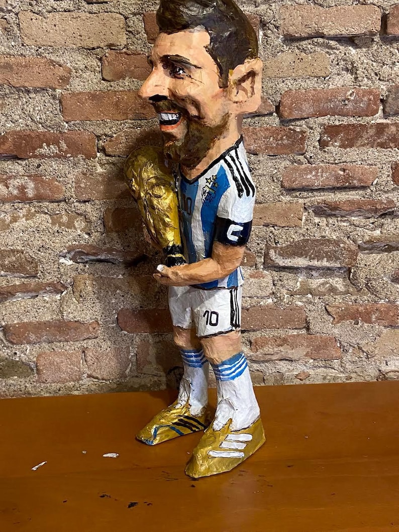 May include: A paper mache figurine of a soccer player holding a gold trophy. The player is wearing a blue and white jersey with the number 10 on the back. The figurine is standing on a wooden surface.
