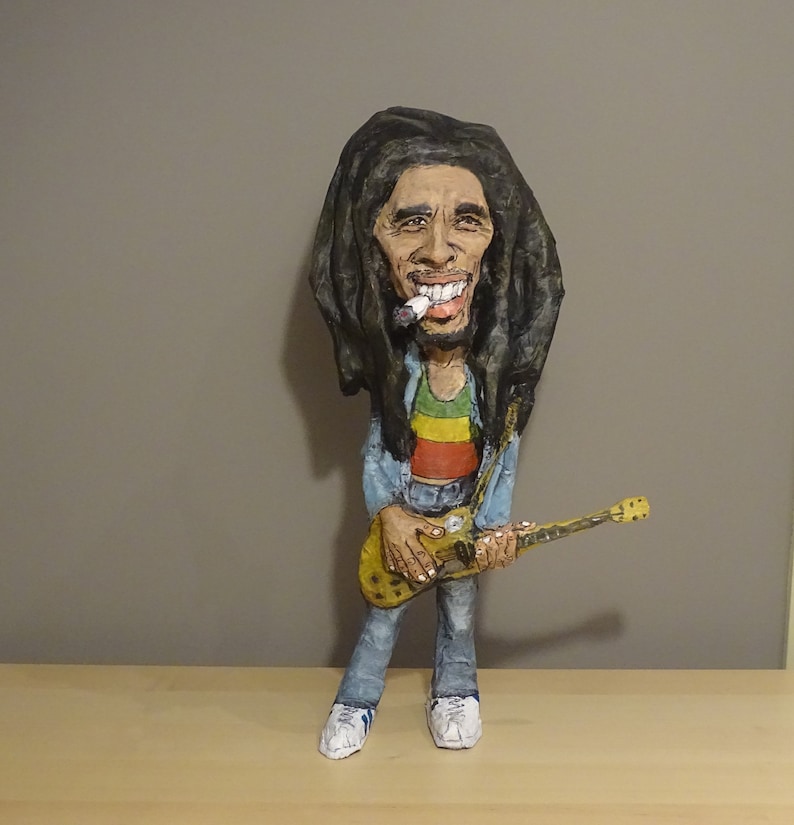 May include: A paper mache figurine of Bob Marley playing a guitar. He is wearing a blue shirt, red, yellow, and green striped shirt, and blue jeans. He has long dreadlocks and is holding a cigarette in his mouth.
