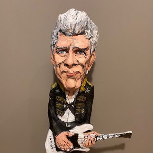 Könnte beinhalten: Eine handgefertigte Figur eines Mannes mit grauem Haar, der eine weiße E-Gitarre spielt. Er trägt eine schwarze und goldene Militärjacke mit Sternen am Kragen.