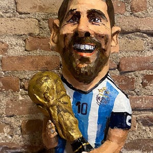 May include: A caricature of a soccer player wearing a blue and white striped jersey with the number 10 on the front. He is holding a gold trophy in his right hand and smiling.