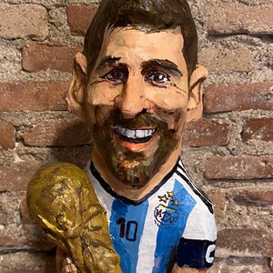 May include: A paper mache figurine of a soccer player holding a golden trophy. The player is wearing a blue and white striped jersey with the number 10 on the front. The jersey has a blue patch with stars and the letters AFA.