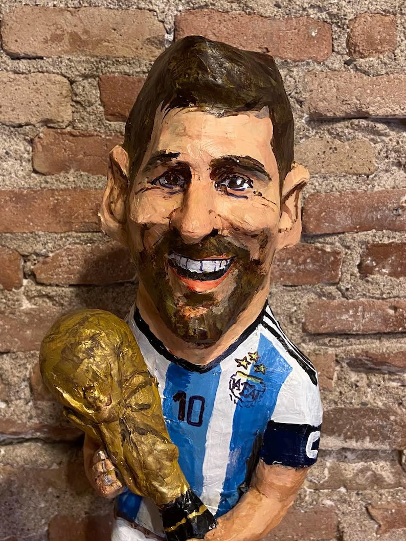 May include: A paper mache figurine of a soccer player holding a golden trophy. The player is wearing a blue and white striped jersey with the number 10 on the front. The jersey has a blue patch with stars and the letters AFA.