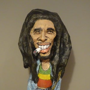 May include: A paper mache sculpture of Bob Marley with long dreadlocks, wearing a blue shirt and a red, yellow, and green striped shirt underneath. He is smiling and holding a cigarette in his mouth.