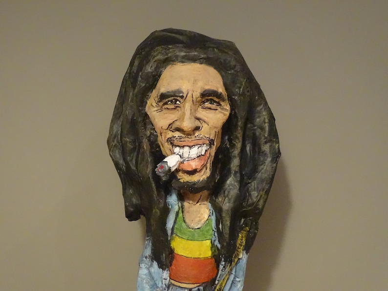 May include: A paper mache sculpture of Bob Marley with long dreadlocks, wearing a blue shirt and a red, yellow, and green striped shirt underneath. He is smiling and holding a cigarette in his mouth.