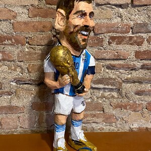 May include: A paper mache figurine of a soccer player holding a gold trophy. The player is wearing a blue and white jersey with the number 10 on it. The figurine is standing on a wooden surface.