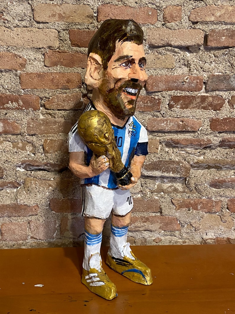 May include: A paper mache figurine of a soccer player holding a gold trophy. The player is wearing a blue and white jersey with the number 10 on it. The figurine is standing on a wooden surface.