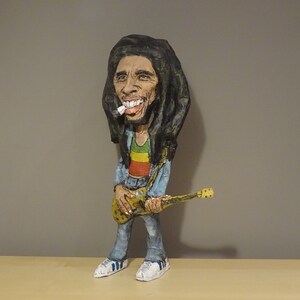 May include: A paper mache figurine of Bob Marley holding a guitar. He is wearing a blue shirt, red, yellow, and green pants, and a black and brown wig.
