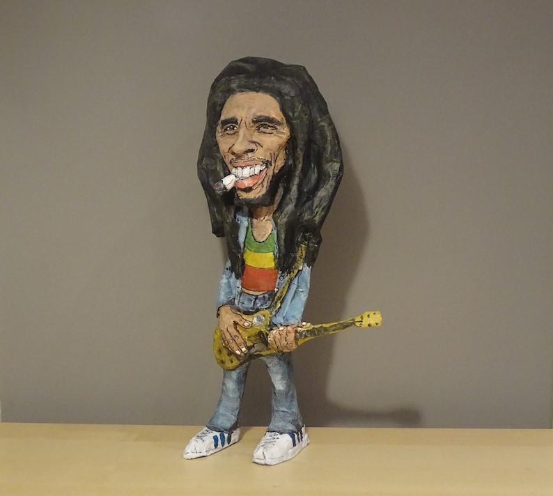May include: A paper mache figurine of Bob Marley holding a guitar. He is wearing a blue shirt, red, yellow, and green pants, and a black and brown wig.