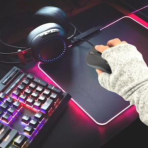 Peut inclure: Un casque de jeu noir avec des anneaux lumineux LED bleus, une souris d'ordinateur noire et un clavier mécanique aux touches rétroéclairées multicolores. Les périphériques sont disposés sur un tapis de souris rectangulaire à bordure LED rose.