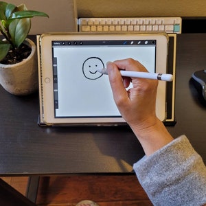 Puede incluir: Una persona utiliza un lápiz digital para dibujar una cara sonriente en una tableta de dibujo digital. La tableta está sobre un escritorio negro con un teclado y un ratón.