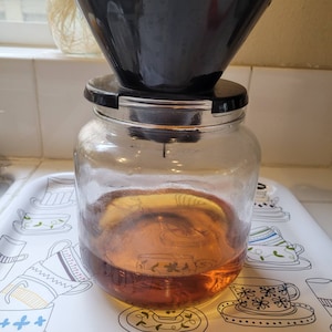 May include: A clear glass jar with a black lid and a black coffee filter sitting on top. The jar is filled with a dark brown liquid.