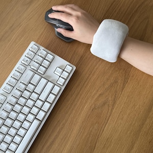 May include: A light gray wrist rest is worn on a person's wrist while using a computer mouse. A white keyboard is in the foreground. The wrist rest appears to be made of a soft, cushioned material. The mouse is dark gray.