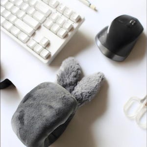 Pode incluir: Uma faixa de orelhas de coelho de pelúcia cinza é o ponto focal desta imagem, cercada por um teclado branco, um mouse ergonômico preto e outros materiais de escritório. A faixa é macia e fofa, com duas orelhas em pé.
