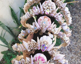 May include: A set of five white and pink seashell crowns with gold accents and iridescent rhinestones. The crowns are stacked on top of each other, creating a cascading effect.
