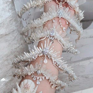 May include: Elaborate white and silver arm jewelry featuring crystal and gem embellishments. The pieces include multiple armbands, bracelets, and a hand piece, all adorned with sparkling details. The design evokes a fantasy or bridal theme.