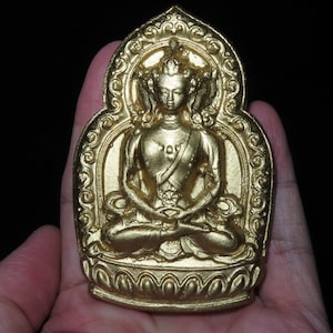 May include: A gold-colored metal pendant depicting a seated Buddha figure with intricate details. The Buddha is surrounded by a decorative frame with floral patterns.