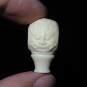 Könnte beinhalten: Eine kleine, weiße, geschnitzte Figur eines Gesichts mit einem breiten Lächeln und einem spitzen Hut. Die Figur ist aus Elfenbein gefertigt und hat ein detailliertes Design.