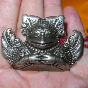 Pode incluir: Um pingente de metal prateado com um relevo detalhado de uma criatura mítica com asas, um bico pontiagudo e mãos juntas sobre o peito. O ornamento apresenta padrões em relevo intrincados e um pequeno cordão vermelho preso ao lado direito.