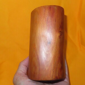 May include: A cylindrical wooden vessel with a smooth, polished finish. The wood displays a warm, reddish-brown colour with visible natural grain patterns, swirls, and small knots throughout the surface, showcasing the organic texture of the material.